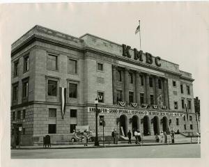 Front of KMBC building exterior at 11th and Central from left side. Grand Reopening sign spanning front archways. Several men and woman in entryway and on sidewalk.